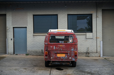 a rusty combi van with no number plate and a sticker 'volksworld 95531510' on a concrete slab with oil stains and behind is the facade of a factoryette with roller doors, windows and a steel door
