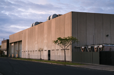 2 sides of a huge factory made out of concrete with a tree and footpath and street in front with big industrial ducts on the roof and a tank on the side and a blue sky with clouds in the background