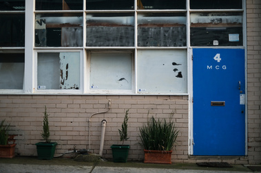 close up of a factoryette facade with a blue door saying '4, MCG' and square  windows with peeling white paint  and a brick wall with pot plants
