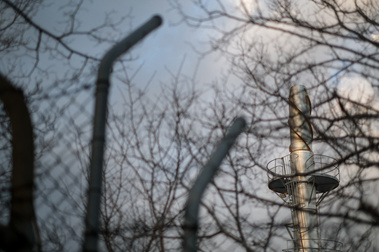 an old high steel mesh fence and winter trees without leaves with a shiny metal industrial  pole shaped large piece of infrastructure and blue sky
