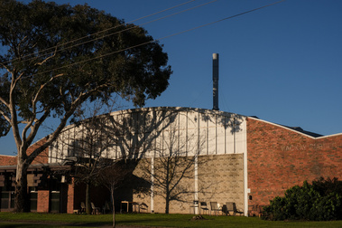 Old red and blonde brick industrial factory with dome shaped roof and metal chimney, blue sky behind and a large gum tree in front causing shadows in the building