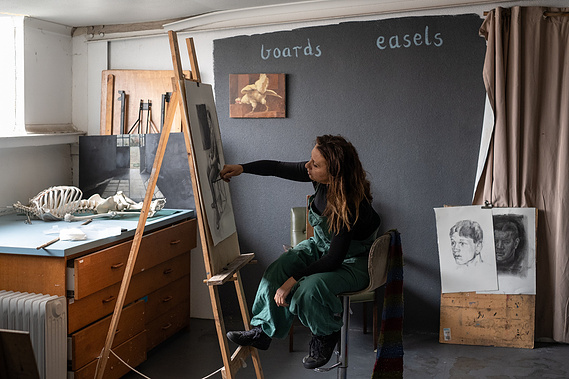 woman sitting painting at an easel in a studio that has a skeleton and paintings and the words 'easel' and 'boards'