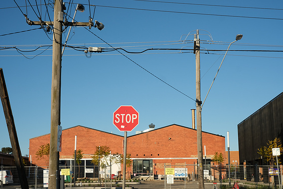 looking past two power poles and power lines, a stop sign and fencing through to an old huge red brick factory with 2 roof peaks and blue sky above 
