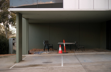 2 carpark spaces with oil stains under a modern office building with three chairs placed randomly with a witches hat