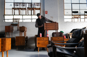 A man oiling a  vintage bedside table surrounded by other pieces of mid century Scandinavian furniture in a sawtooth roof factoryette with painted white floors and walls and steel sash windows behind him
