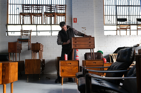 A man oiling a  vintage bedside table surrounded by other pieces of mid century Scandinavian furniture in a sawtooth roof factoryette with painted white floors and walls and steel sash windows behind him
