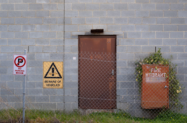 a grey besser brick wall with a rusted steel door, a 'no parking' sign, a 'beware of vehicles' sign, and a steel box with words 'fire hydrant' and a weed growing over it, all seen through a steel mesh fence