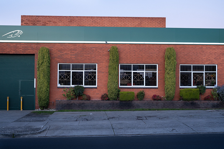 a factory  facade made of red brick and white windows  with a painted green stripe and white jaguar with green roller door and sculpted hedges 