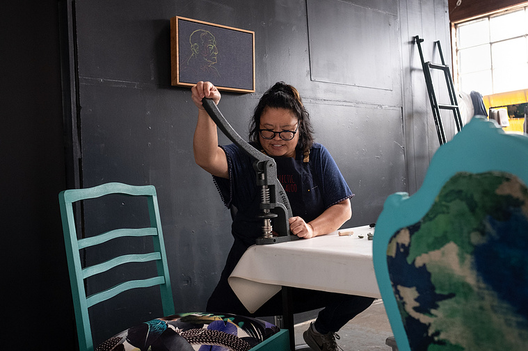 upholsterer sitting at a work table using a button press  with 2 upholstered chairs, a painting and a ladder in her factoryette with a black wall 