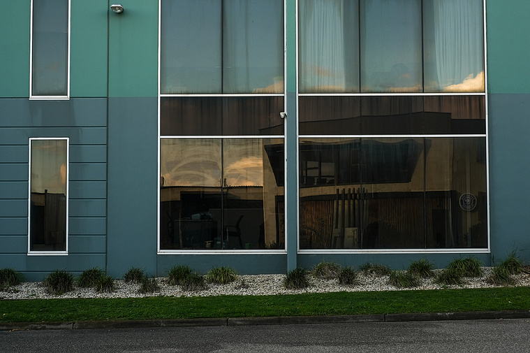 Office building concrete facade painted blue and green with big windows reflecting another office building and plants in front