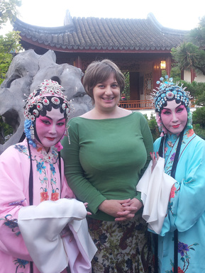 At a Chinese opera performance in Vancouver's Dr. Sun Yat Sen Classical Chinese Garden, the author is pictured with short hair and a green top between 2 of the performers in their opera costumes.