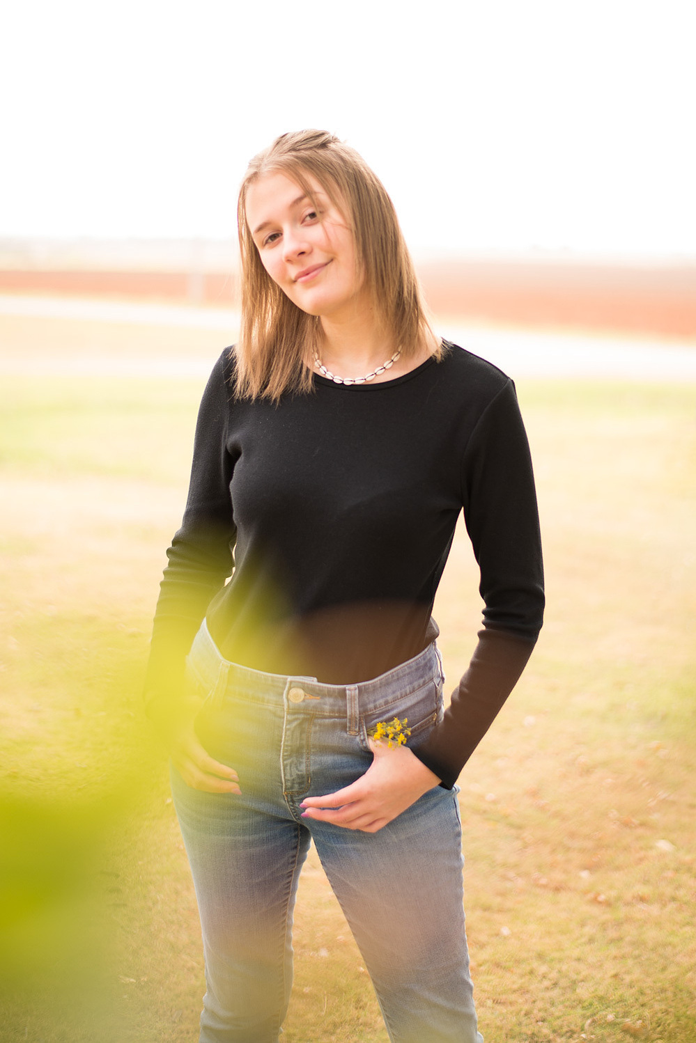 Portrait of a teenage girl in a black t-shirt and jeans standing in a yard smiling with green blurry leaves in the foreground