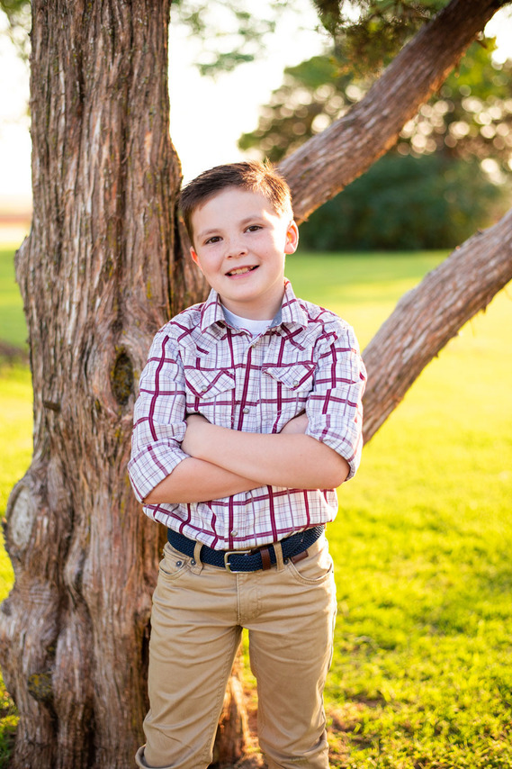 A little boy stands in front of a tree with his arms crossed and smiles in his back to school photo session in the country in Oklahoma