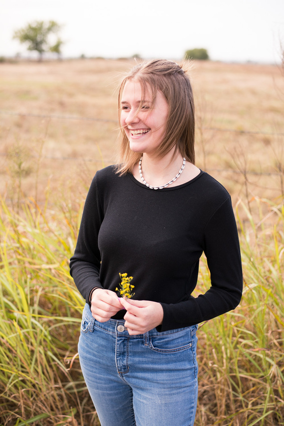 Portrait of a teenage girl in a black t-shirt and jeans in front of a field, holding a small bouquet of yellow wildflowers in her fingers, looking off to the side with a pretty smile 