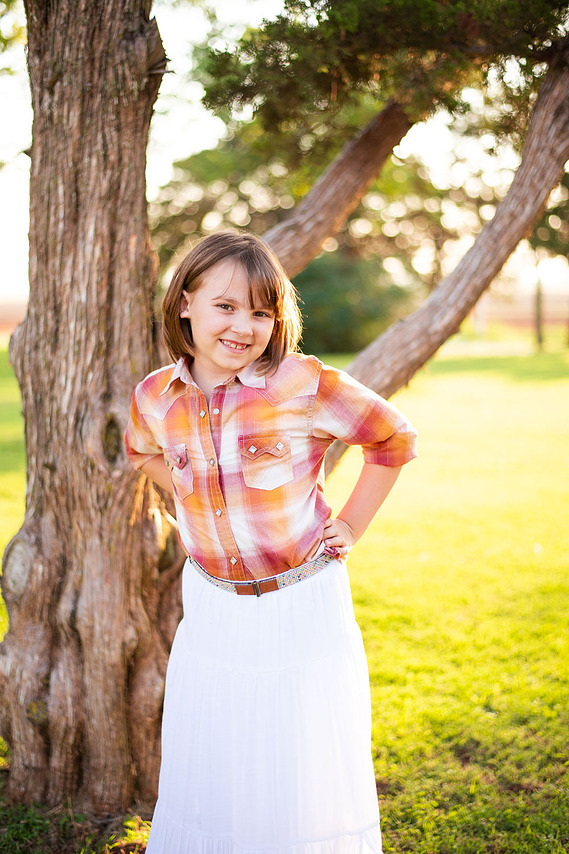 A little girl smiles with her hand on her hip in front of a tree at her back to school photo session in the country in Oklahoma