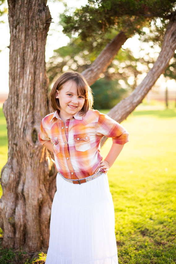 A little girl smiles with her hand on her hip in front of a tree at her back to school photo session in the country in Oklahoma