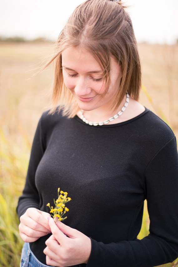 Portrait of a teenage girl in a black t-shirt and jeans in front of a field, holding a small bouquet of yellow wildflowers in her fingers, looking down to admire them.
