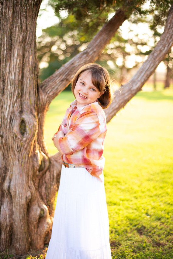 A little girl smiles with her arms crossed in front of a tree at her back to school photo session in the country in Oklahoma