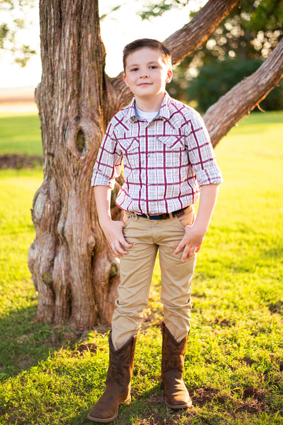A little boy stands in front of a tree with his hands in his pockets and smiles in his back to school photo session in the country in Oklahoma