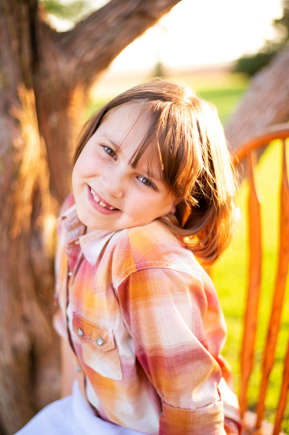A little girl smiles while sitting in a chair in front of a tree at her back to school photo session in the country in Oklahoma