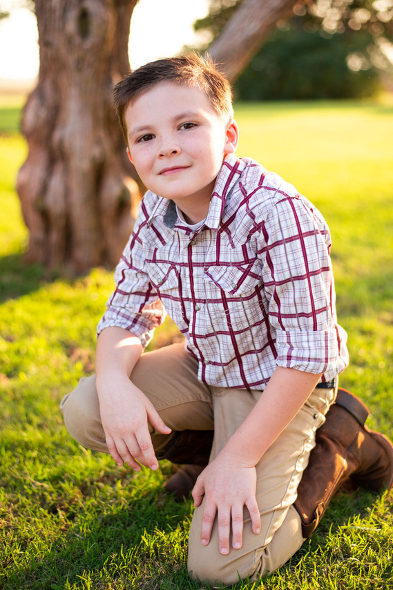 A little boy kneels in a yard in front of a tree in his back to school photo session in the country in Oklahoma