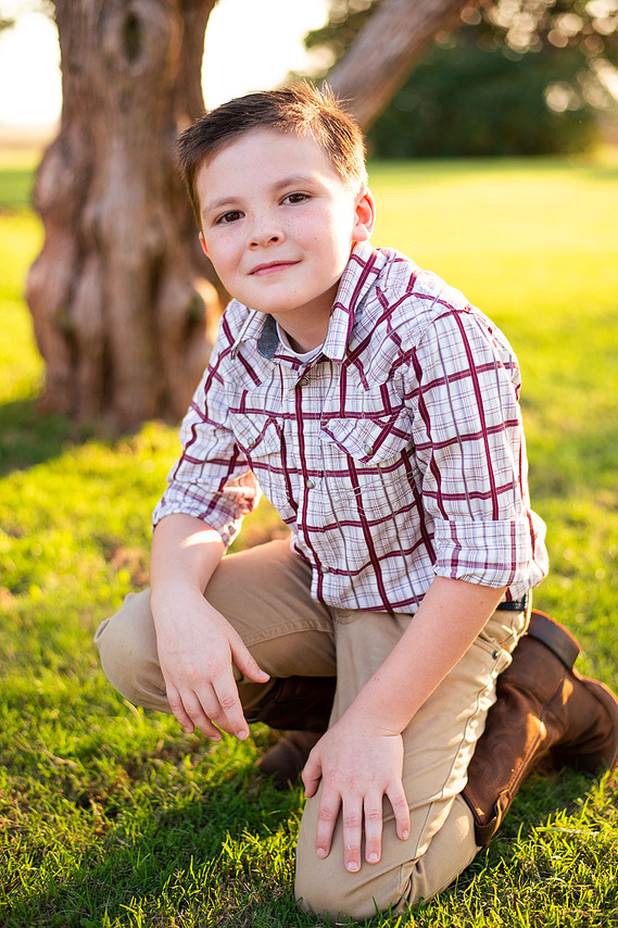A little boy kneels in a yard in front of a tree in his back to school photo session in the country in Oklahoma
