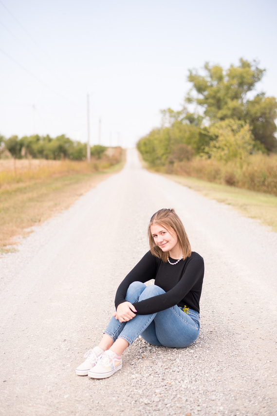 Portrait of a teenage girl in a black t-shirt and jeans sitting in the middle of a dirt road with her hands around her knees smiling at the camera