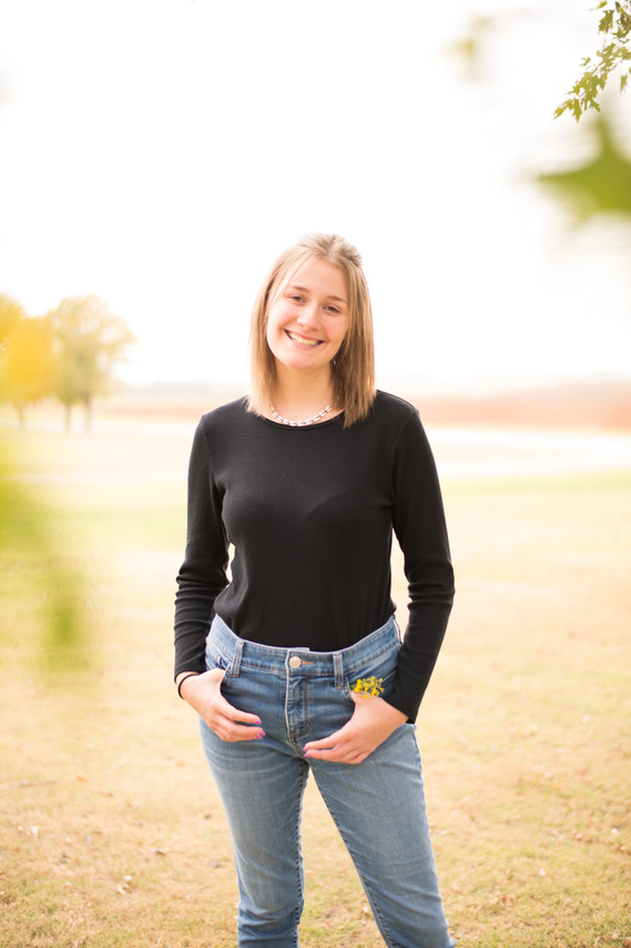 Portrait of a teenage girl in a black t-shirt and jeans standing in a yard smiling with green blurry leaves in the foreground