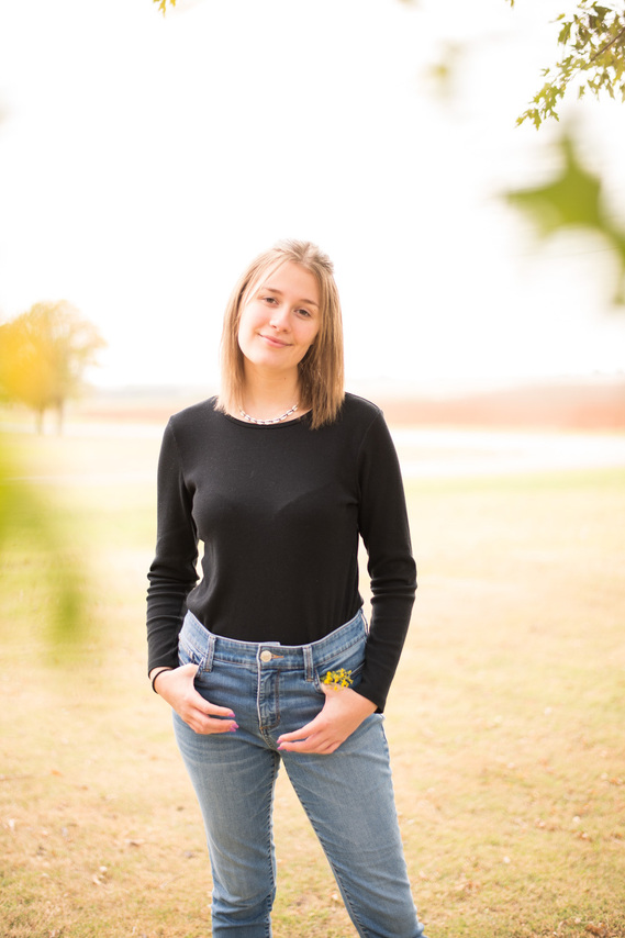 Portrait of a teenage girl in a black t-shirt and jeans standing in a yard smiling with green blurry leaves in the foreground