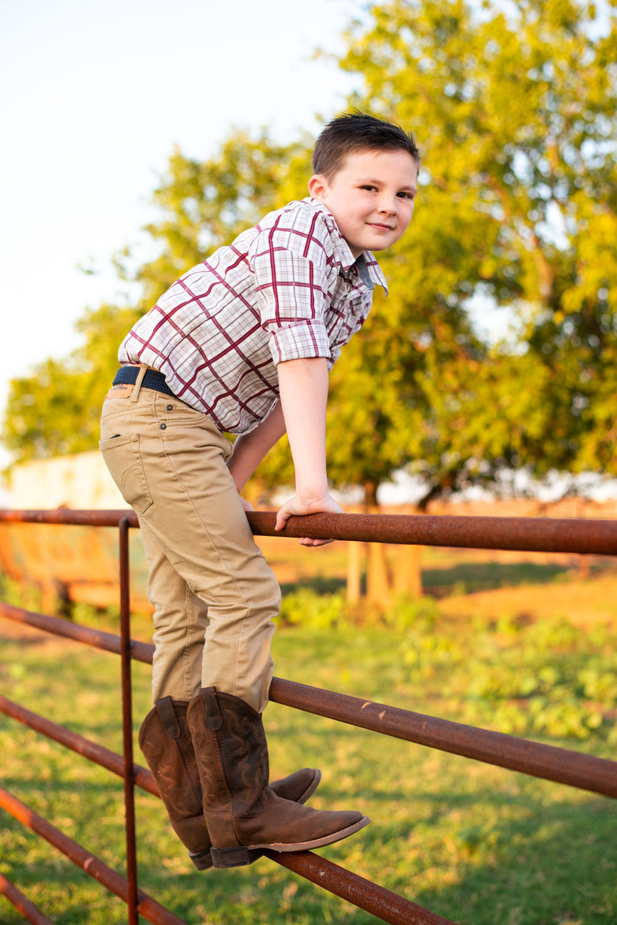 A little boy stands on a metal cattle fence in his cowboy boots in his back to school photo session in the country in Oklahoma