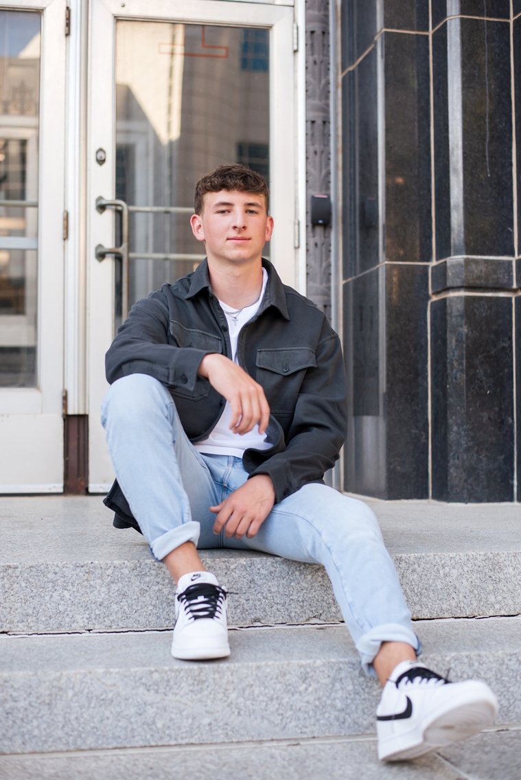 high school senior boy wearing a charcoal gray jacket and blue jeans and white sneakers sits on gray steps in front of entrance doors of a building in downtown OKC