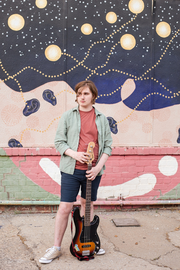 senior boy wearing a green shirt and blue shorts stands holding his bass guitar in front of him. standing in front of a wall mural in midtown Oklahoma City