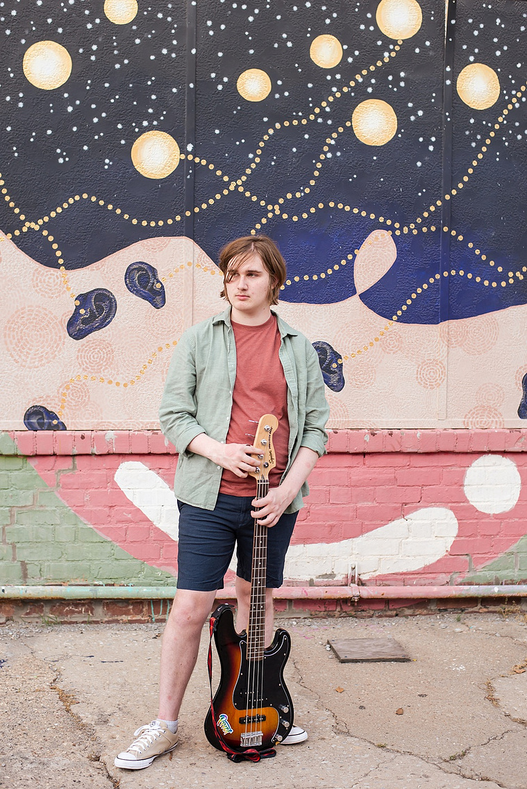 senior boy wearing a green shirt and blue shorts stands holding his bass guitar in front of him. standing in front of a wall mural in midtown Oklahoma City