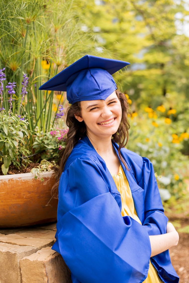 high school senior girl with long brown hair wearing a yellow top and jeans in her blue cap and gown leans back on a ledge with flowers around her at Myriad Gardens in Oklahoma City