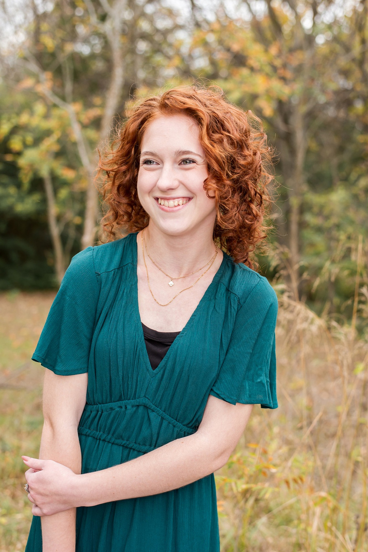 senior girl with red hair wearing teal dress poses and smiles prettily standing in a field with gold grass and fall trees behind her at Sutton Park in Norman, Oklahoma