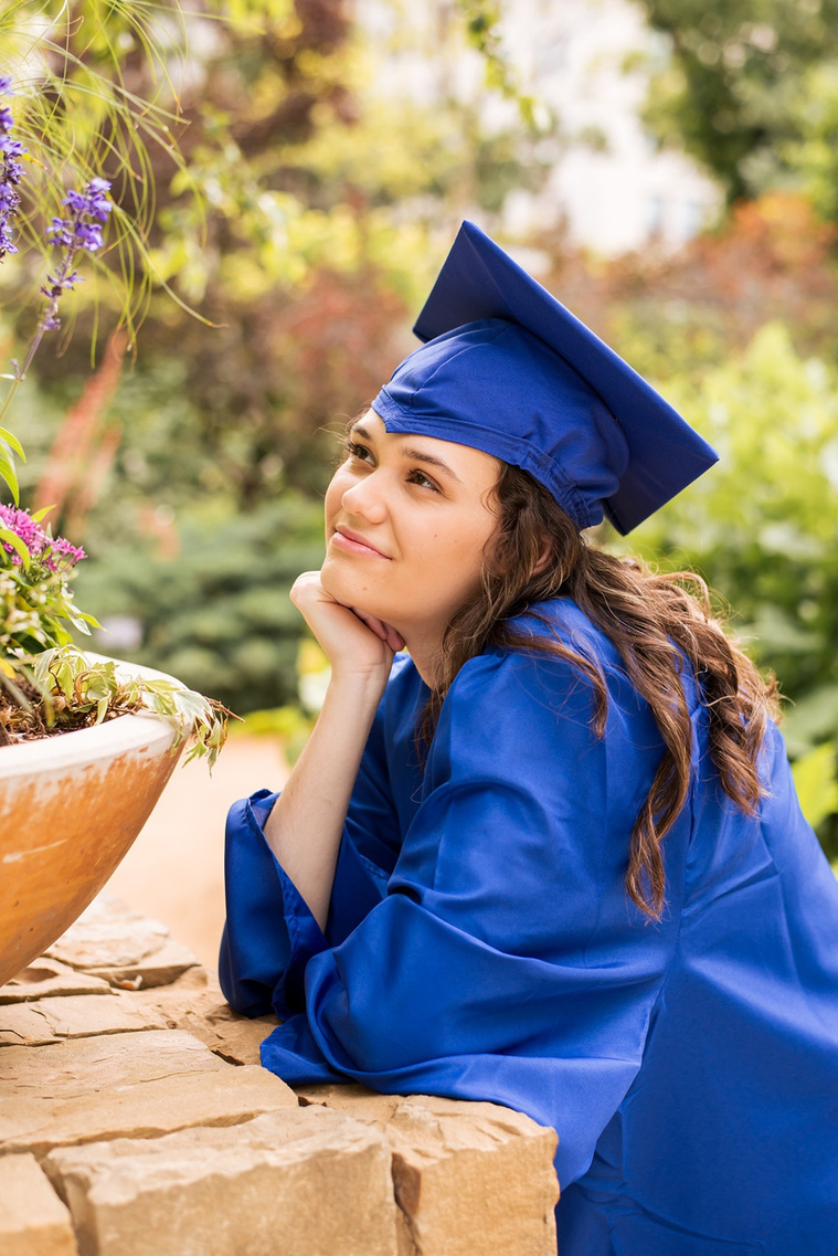 high school senior girl with long brown hair wearing a yellow top and jeans and a blue cap and gown leans on an elbow with chin in hand looking up at a pot of flowers at Myriad Gardens in Oklahoma City