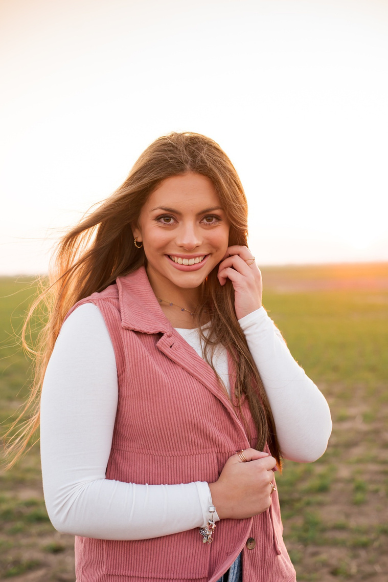 portrait of a beautiful senior girl with long hair wearing a pink vest smiling with one hand in her hair standing in front of a winter wheat field in the country near Oklahoma City
