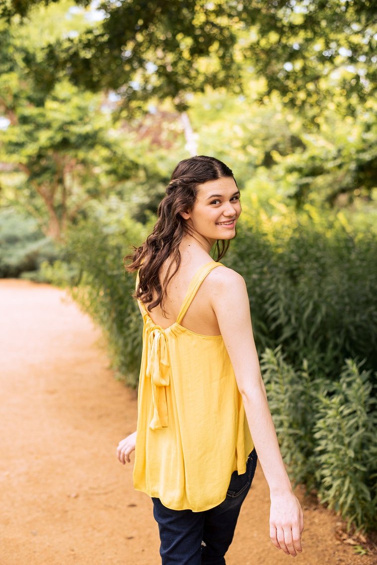 high school senior girl with long brown hair wearing a yellow top and jeans looks over her shoulder smiling while she walks away on a path at Myriad Gardens in Oklahoma City