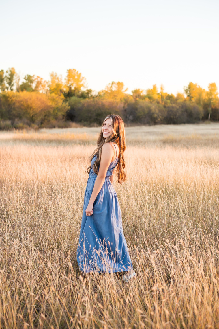 senior girl with long brown hair wearing a blue sundress smiles prettily over her shoulder in a country field in the fall with trees behind her near Oklahoma City