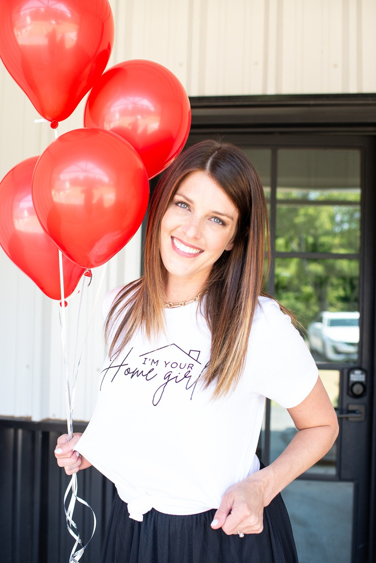 Brand photo shoot of realtor in OKC. She stands by front door holding red balloons and smiling highlighting her t-shirt with a witty saying