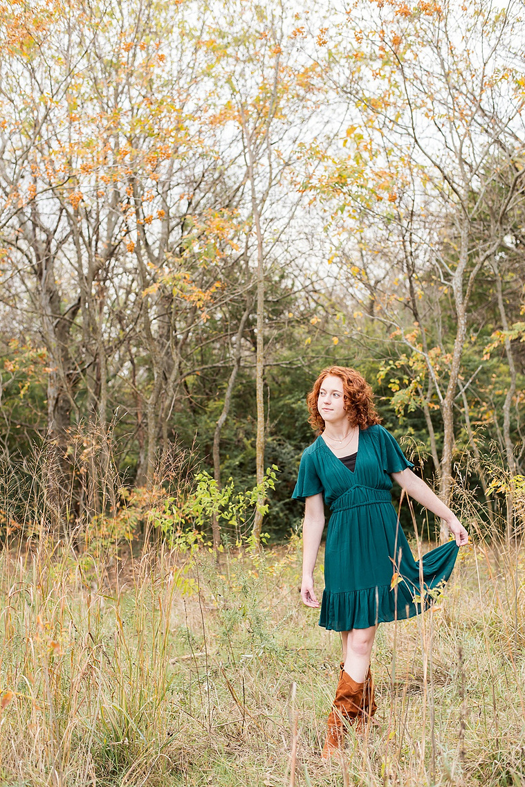 senior girl with red hair wearing teal dress holds skirt out in a dancers pose and looks off standing in a field with gold grass and fall trees behind her at Sutton Park in Norman, Oklahoma