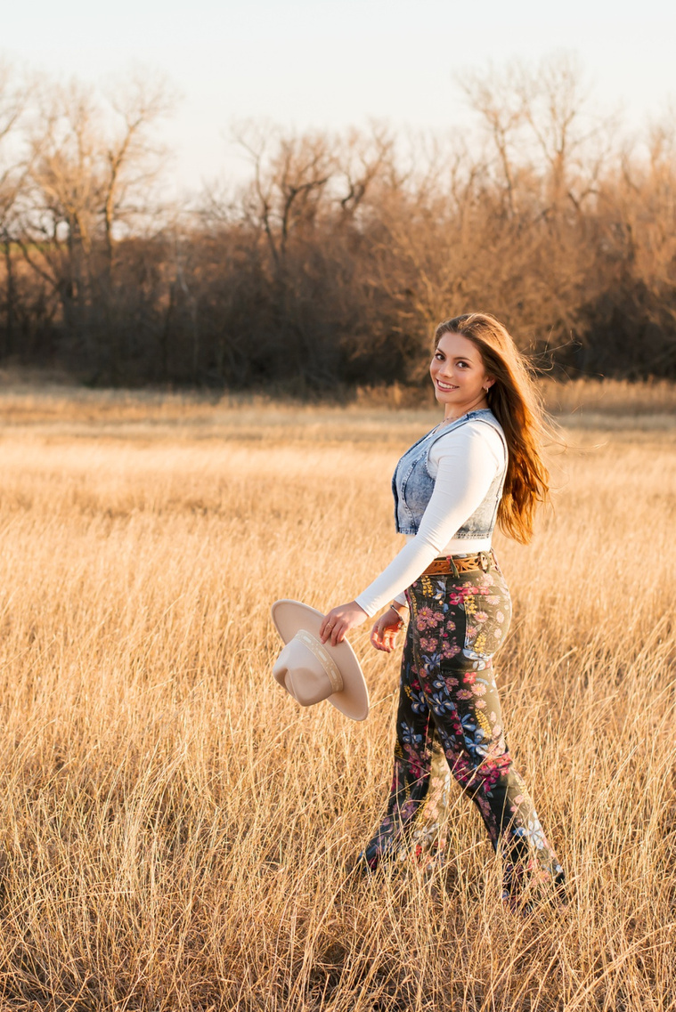 high school senior girl with long brown hair wearing cowboy boots, Wranglers, a denim vest and western hat smiles over her shoulder as she walks through a field of golden grass in the country near OKC