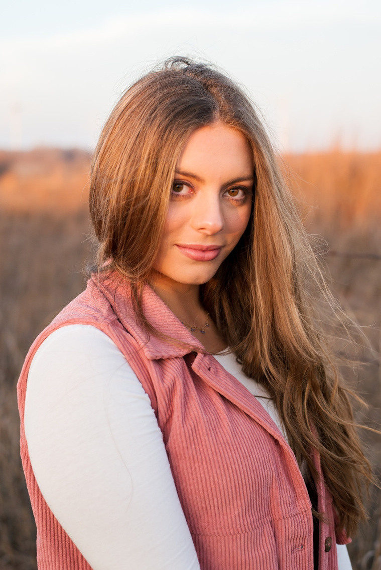 portrait of a beautiful senior girl with long hair wearing a pink vest looking seriously at camera standing in front of country fields in Oklahoma