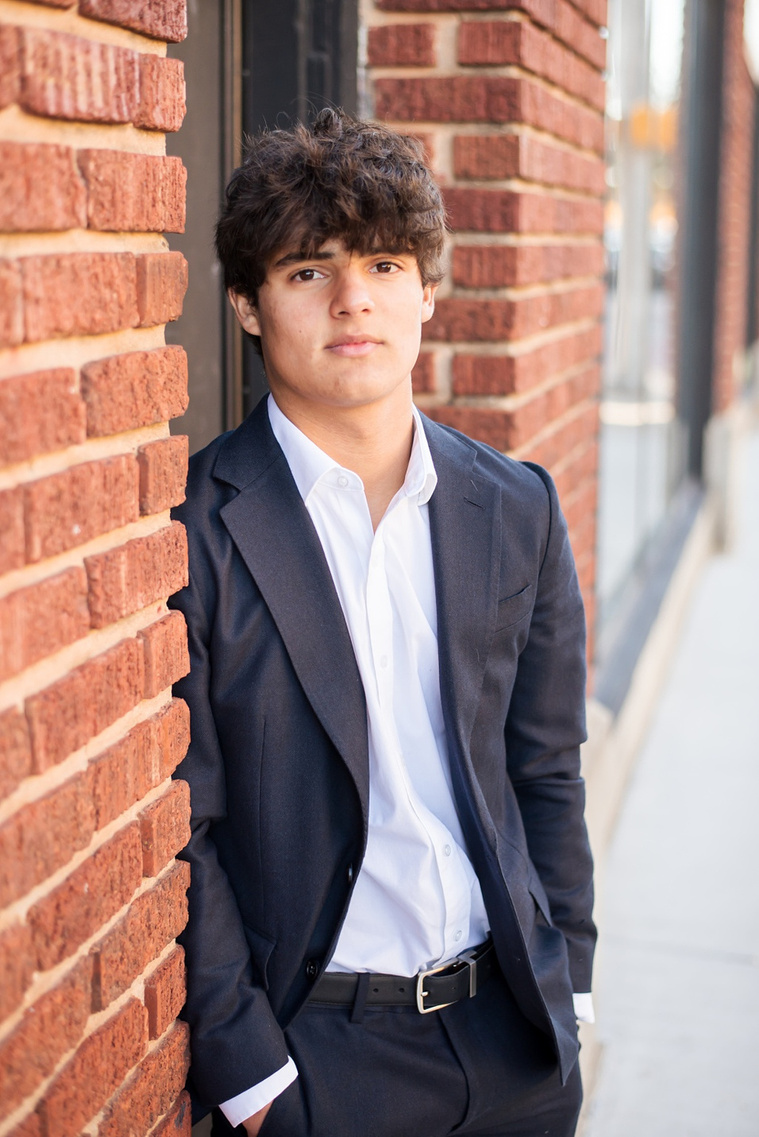 portrait of a hispanic high school senior boy wearing a dark blue suit and white shirt leaning casually against a brick wall and looking at camera with no smile in Automobile Alley in Oklahoma City
