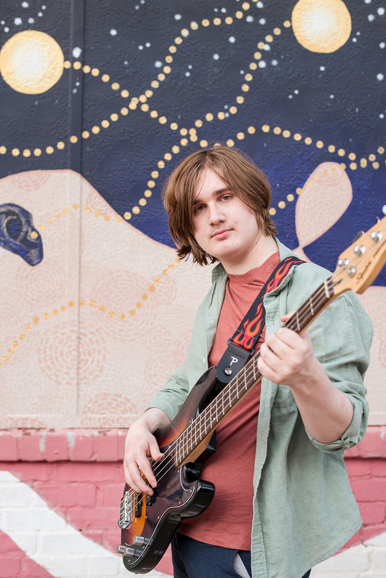 senior boy wearing a button down shirt and blue shorts holds his bass guitar like he's playing and looks at camera, in front of a colorful wall mural in midtown Oklahoma City