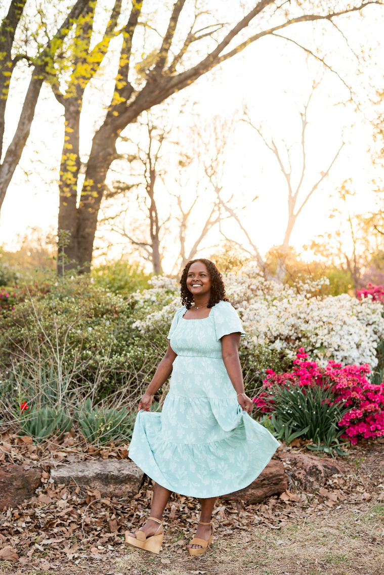 senior girl wearing a dress smiles and  holds the edge of her skirt out as she looks up  among the flowers and trees of Will Rogers Gardens in OKC