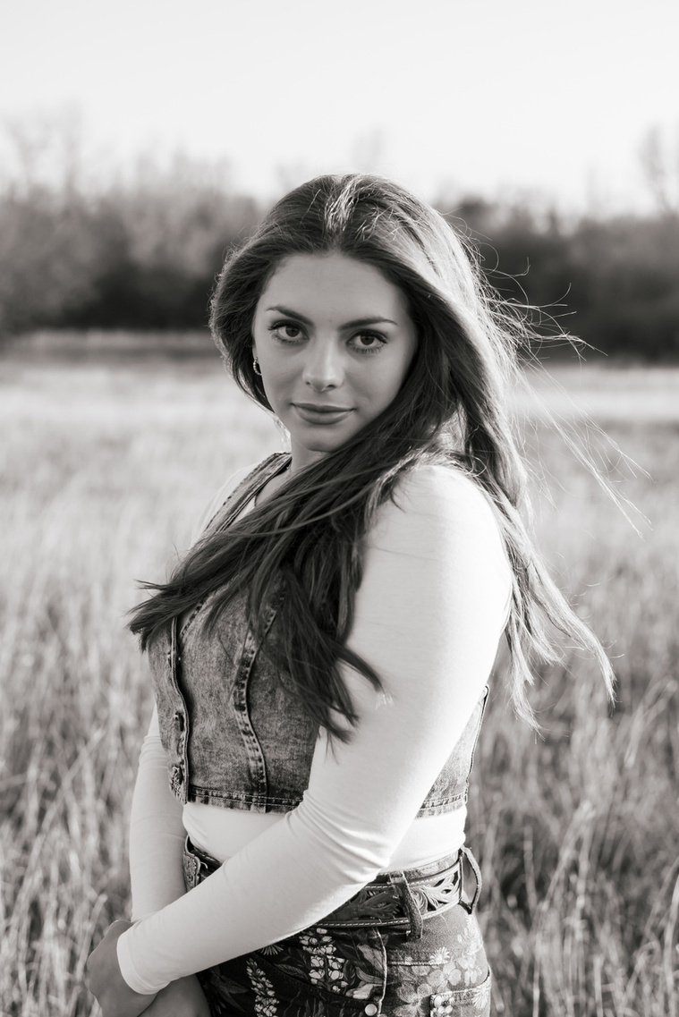 black and white photo of a high school senior girl with long brown hair wearing a denim vest and western jeans looks seriously at the camera while she stands in a country field near OKC