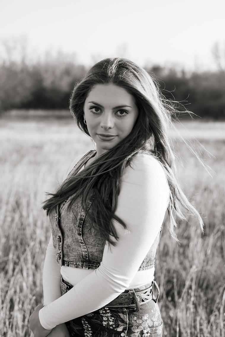 black and white photo of a high school senior girl with long brown hair wearing a denim vest and western jeans looks seriously at the camera while she stands in a country field near OKC