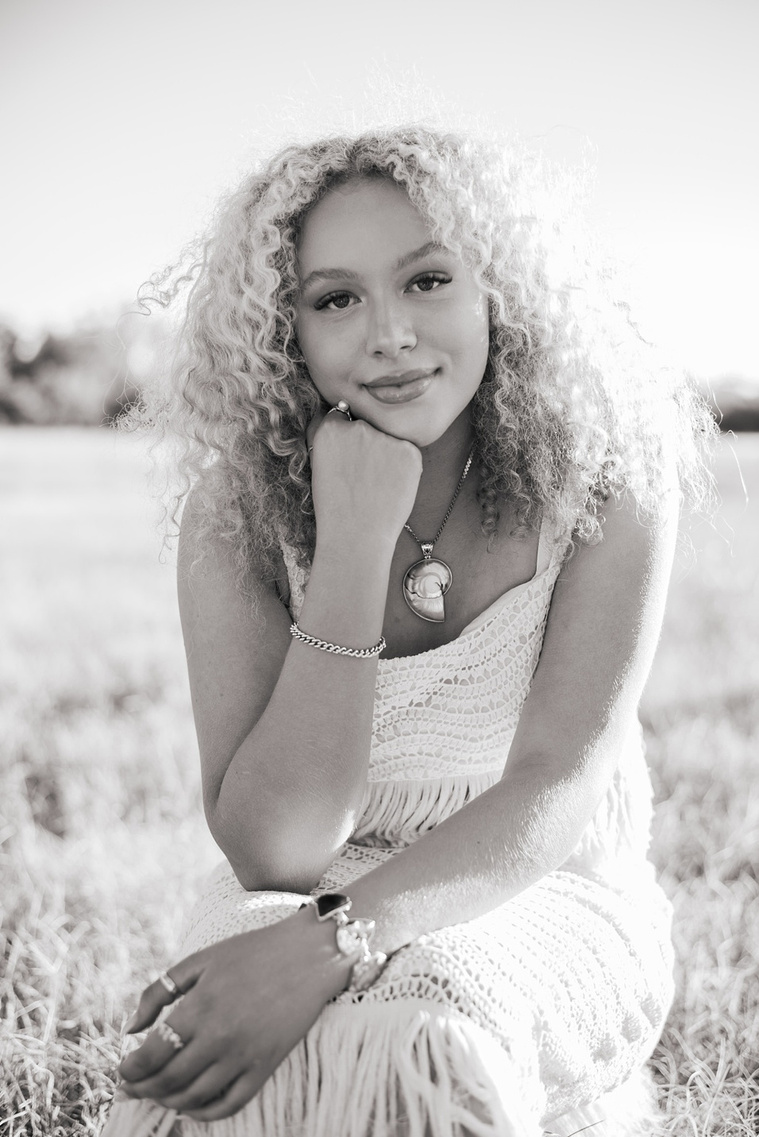 black and white portrait of a high school senior girl with blonde curly hair and blue eyes sitting with her hand on chin and softly smiling in a field at Martin Nature Park in Oklahoma City