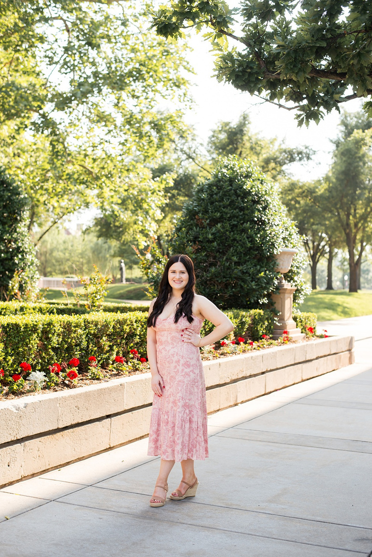college senior girl wearing a light pink floral sundress with long dark hair stands with one hand on waist in front of a flower bed and hedges with trees around her on a sidewalk on OU campus in Norman